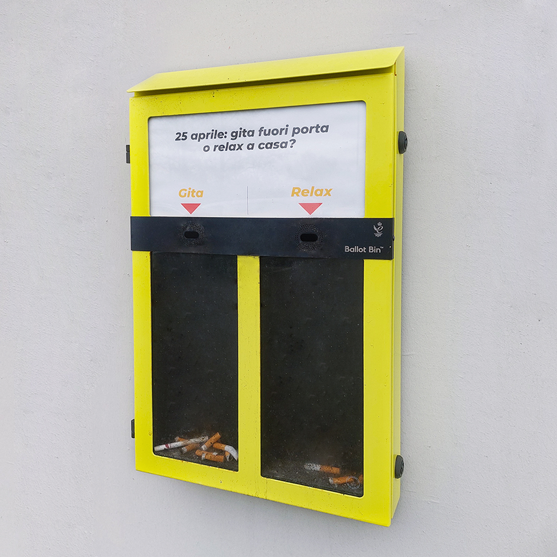 Contenitore giallo Ballot Bin per mozziconi, montato su una parete bianca., all'esterno dell'azienda Satech. Il pannello frontale presenta un sondaggio.