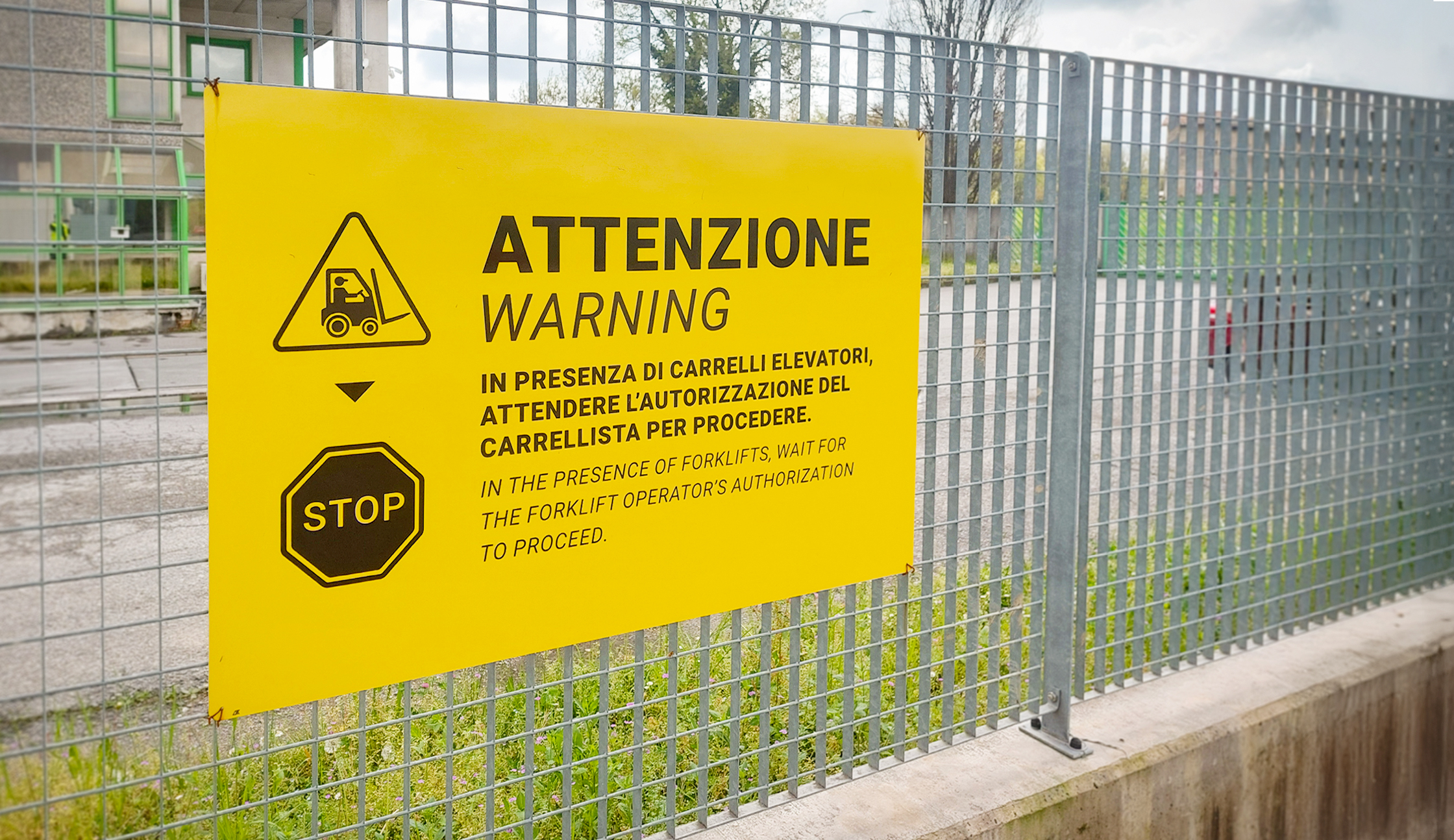 Cartello giallo di avviso per trasportatori, montato su rete in acciaio all’esterno di Satech. Segnala il transito di carrelli elevatori e invita ad attendere l’autorizzazione prima di procedere. Testo in italiano e inglese.