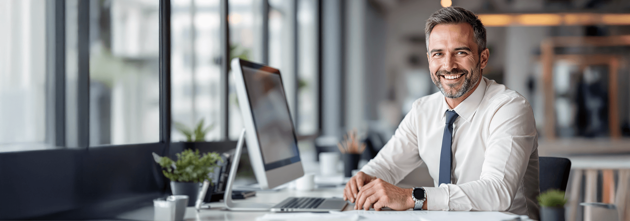 Impiegato seduto alla scrivania sorride verso l’obiettivo. Davanti a lui un computer e alcune piantine; sullo sfondo, una finestra e l’ambiente ufficio.
