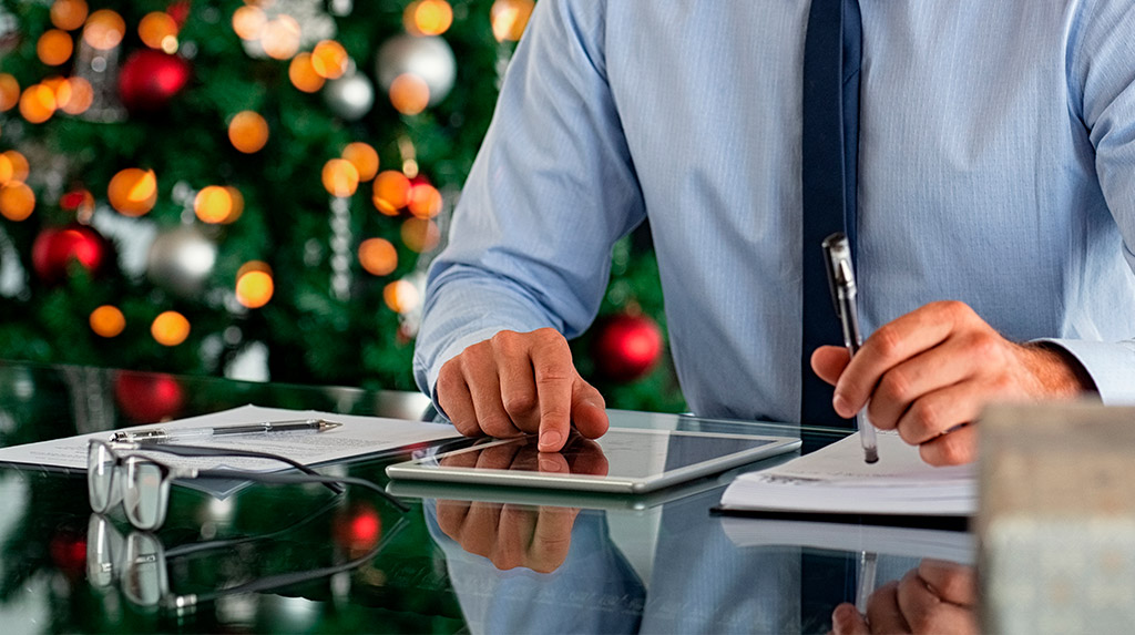 Professionista al lavoro su tablet e documenti durante un’attività di pianificazione, con albero di Natale sullo sfondo.